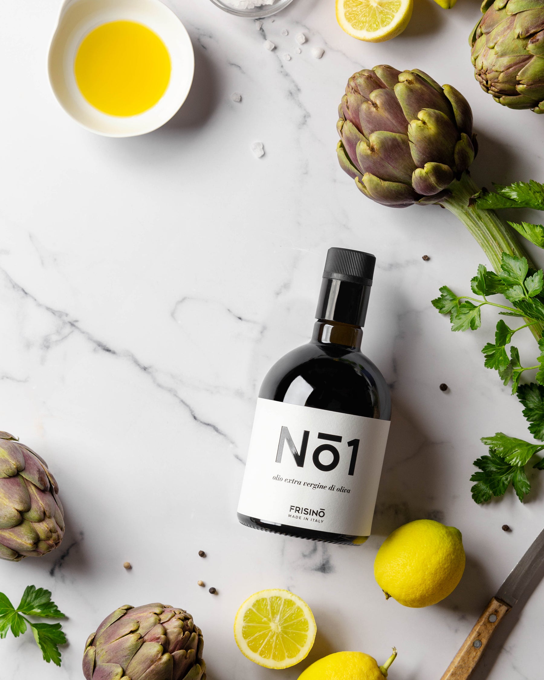 Frisino N°1 Peranzana extra virgin olive oil bottle on marble with artichokes, lemons, parsley and oil bowl