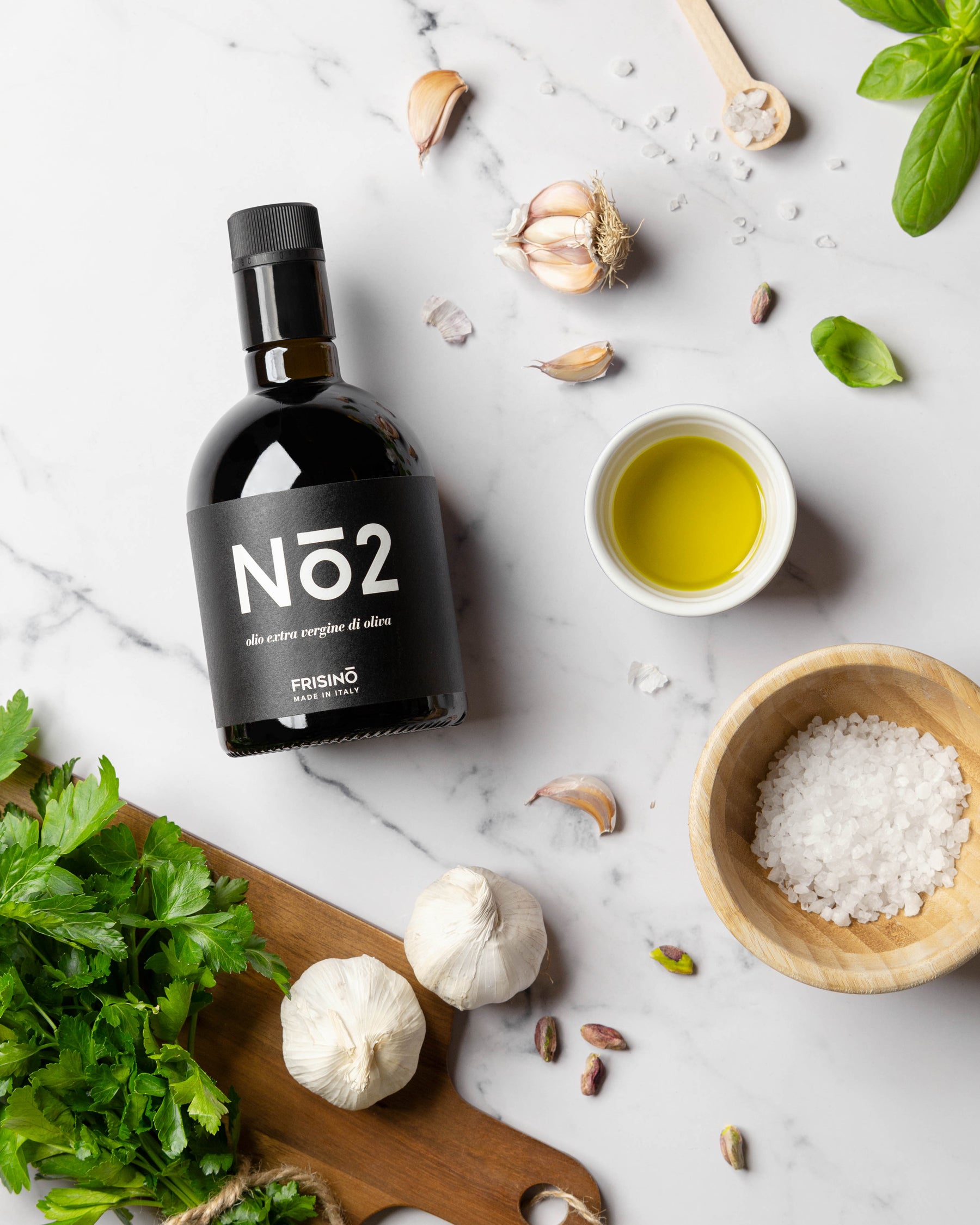 Frisino N°2 extra virgin olive oil bottle on marble with garlic, basil, coarse salt and a bowl of golden oil
