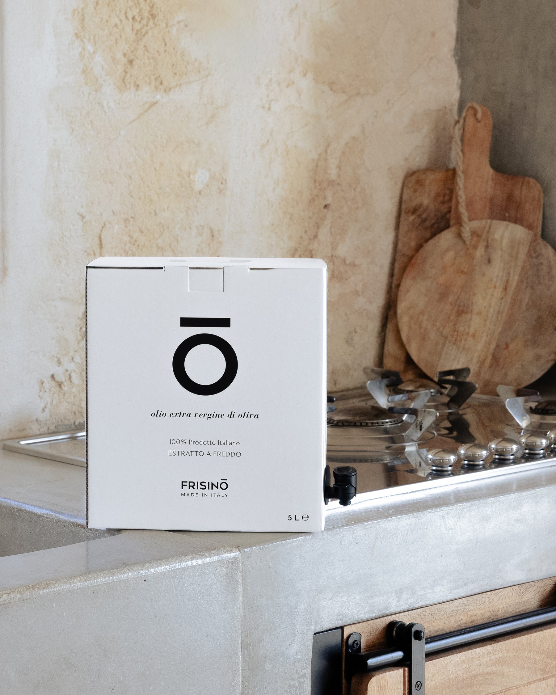Frisino 5 L bag-in-box of Peranzana extra virgin olive oil with tap, on a kitchen counter beside a gas hob and wooden cutting boards