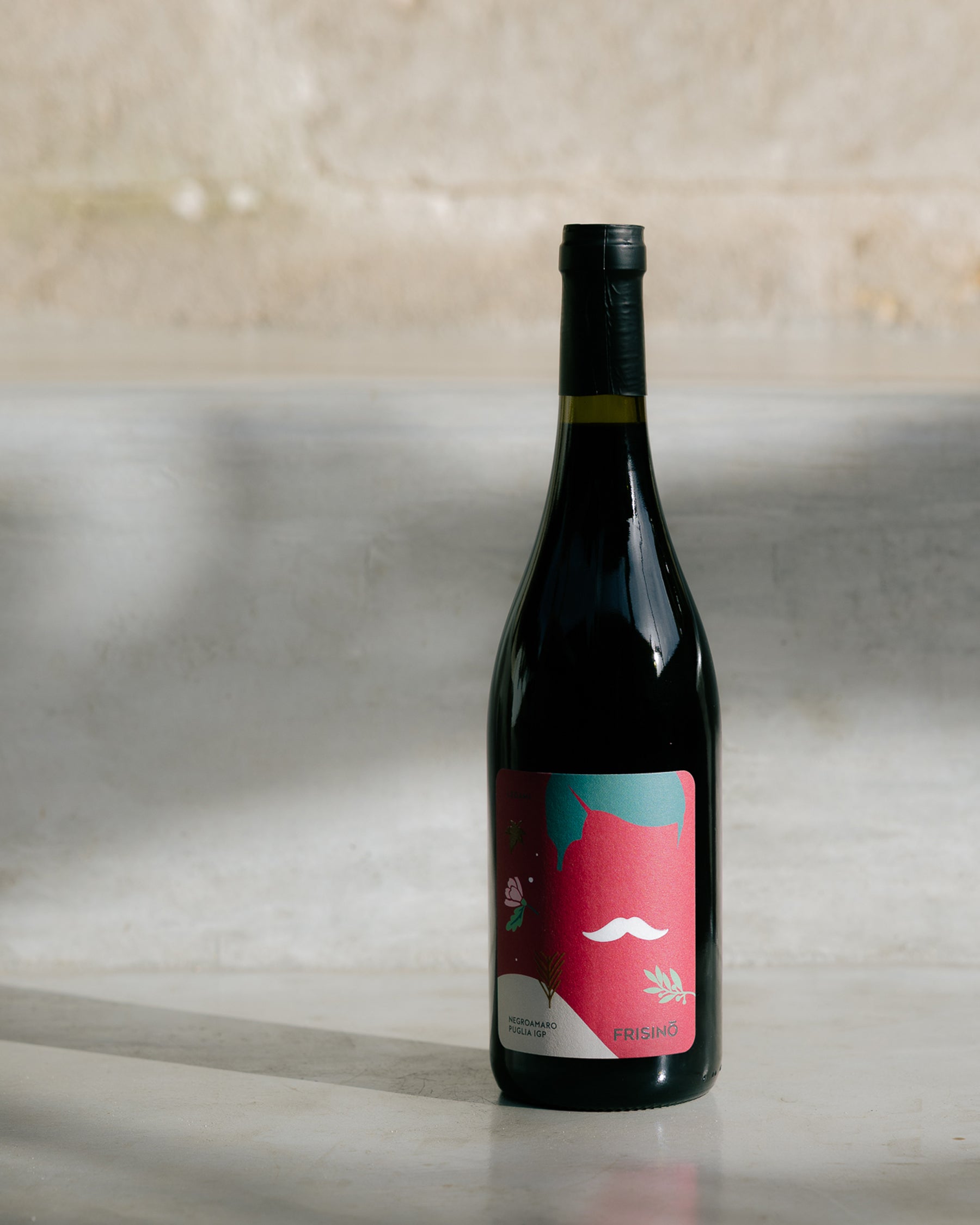 Bottle of Legàmi Negroamaro Puglia IGP by Frisino with pink label and white moustache, on a light concrete background