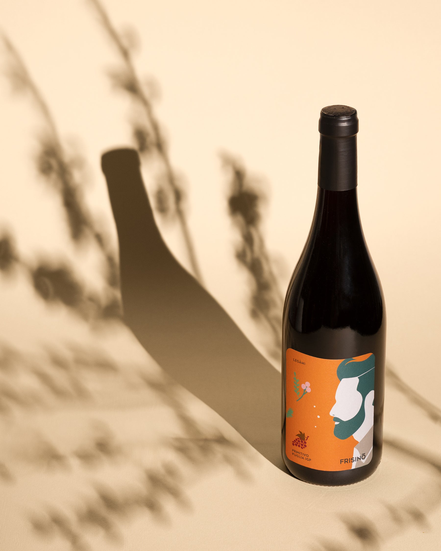 Bottle of Frisino Legàmi Primitivo Puglia IGP with orange label and stylized face, branch shadows on beige backdrop