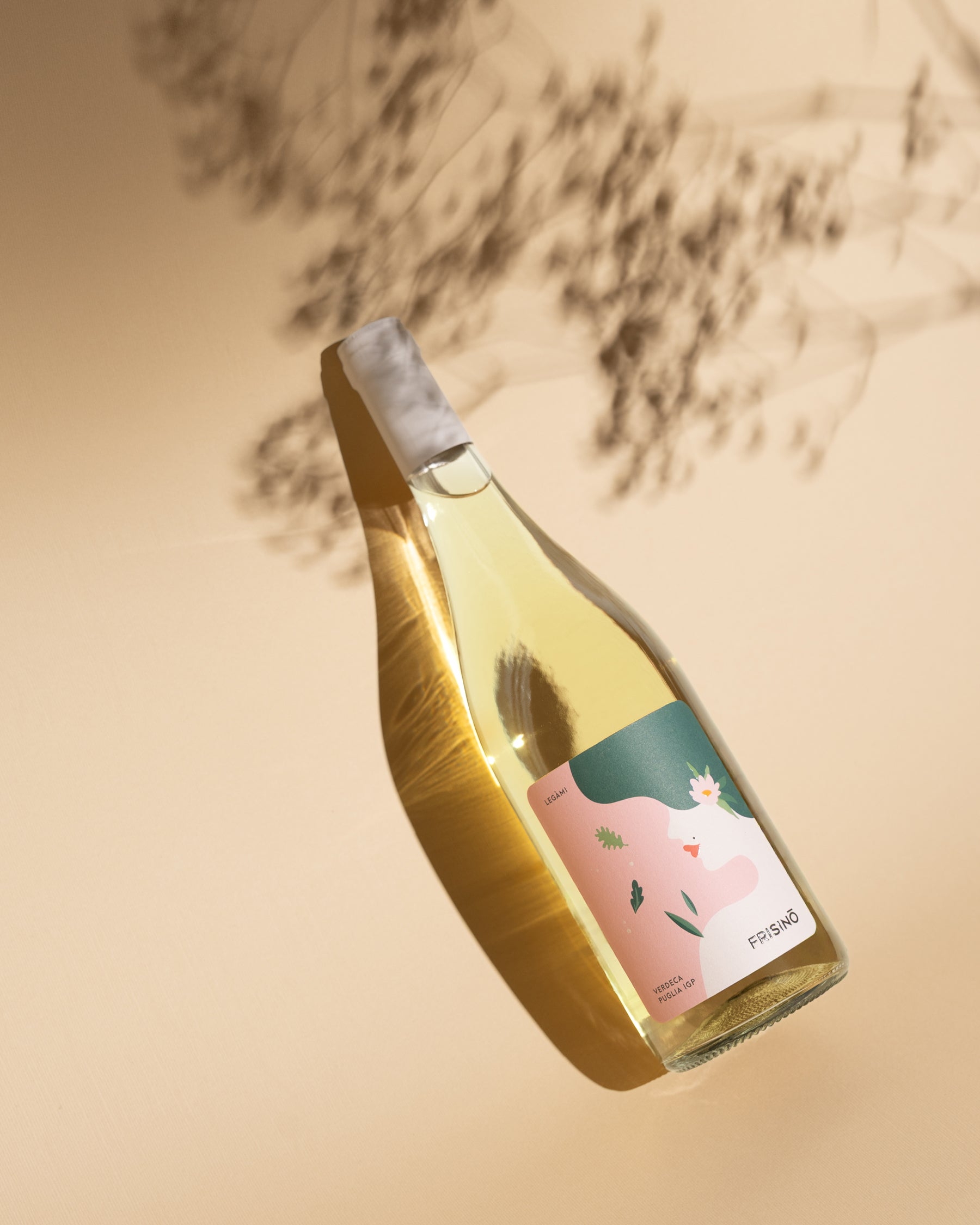 Bottle of Frisino Legàmi Verdeca Puglia IGP with illustrated pink label, resting on a beige surface amid dried branch shadows