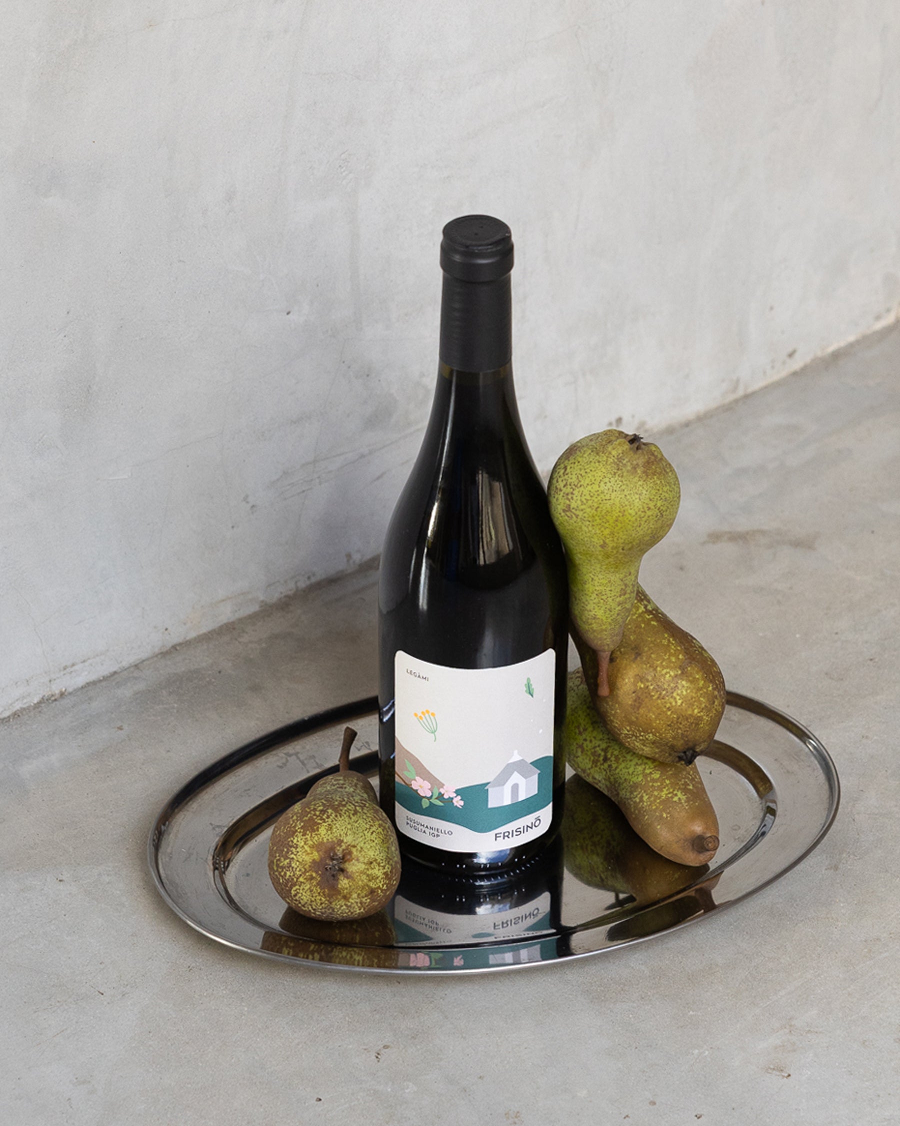 Bottle of Frisino Legàmi Susumaniello Puglia IGP on a silver tray with fresh pears against a concrete backdrop